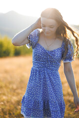 Young woman in a blue floral dress walking through a sunlit meadow during summer