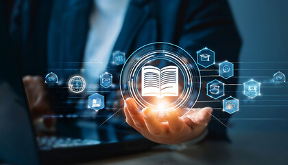 Digital education concept with a glowing open book icon surrounded by virtual learning symbols, displayed above a hand with a blurred laptop and dark background.

