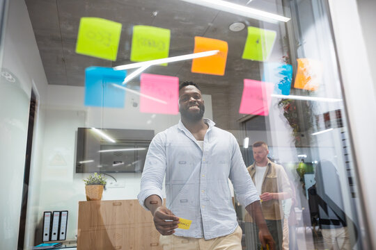 Happy african american manager organizing tasks using colorful sticky notes on a glass wall in a modern coworking space