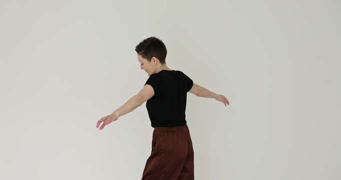 Woman with pixie haircut dances spinning around on white background. Female dancer with spread arms moves gracefully across room slow motion