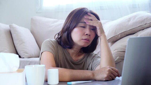 Tired and sick Asian woman working remotely from home while suffering from a cough and illness using her laptop ,concept of remote work during illness, stress on health, being unwell at home