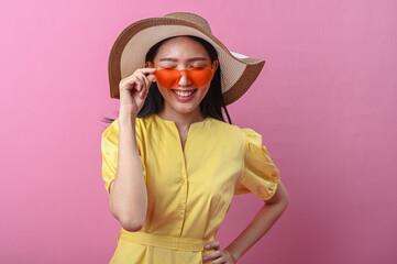 Asian woman in a yellow dress and wide-brimmed hat smiling playfully while holding orange...