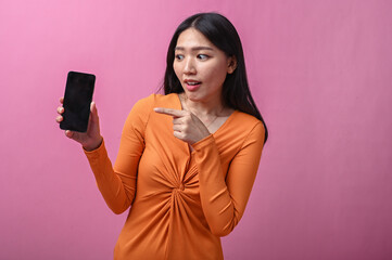 Asian woman with long black hair in orange dress smiling and pointing at smartphone screen with...