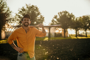Farmer smiling and holding pitchfork in cultivated field at sunset