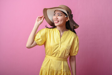 Asian woman in a yellow dress and wide-brim hat smiling softly while touching her hat, posing...