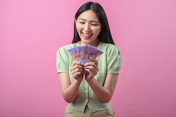 Asian woman in green top smiling happily while holding a fan of purple banknotes in both hands,...