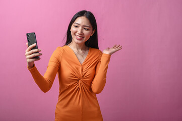 Asian woman with long black hair in orange dress smiling and waving while holding smartphone for a...