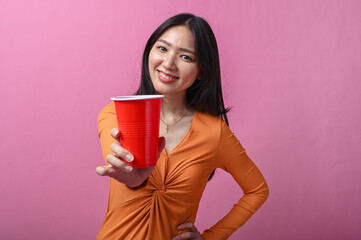 Asian woman with long black hair in orange dress smiling while extending a red plastic cup toward...