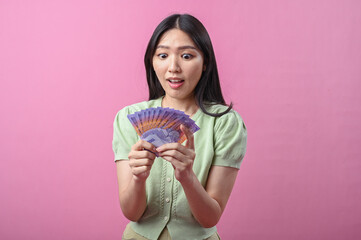Asian woman looking surprised and excited while holding a fan of Indonesian rupiah banknotes,...