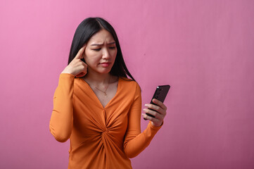Asian woman with long black hair wearing orange dress looking confused while holding smartphone in...