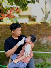 A Father wearing medical gloves holds his sick child and gives him nebulizer therapy in an outdoor...