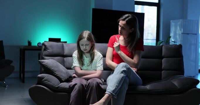 Woman scolds young girl with arms crossed and sullen expression sitting on sofa. Disciplinary moment and difficult conversation at home slow motion