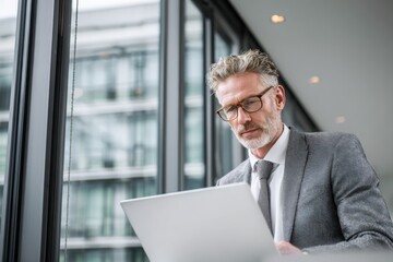 Fototapeta premium Mature professional man using laptop in modern office lounge, portraying remote work, business confidence, and digital productivity.