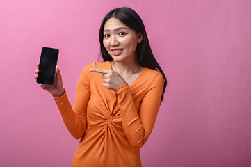 Asian woman with long black hair in orange dress smiling and pointing at smartphone screen with...