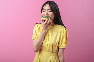 Asian woman in a yellow dress about to bite a green-sprinkled donut while looking at the camera,...