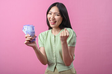 Asian woman smiling confidently while raising her fist and holding a stack of Indonesian rupiah...