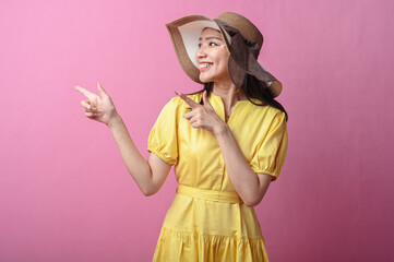 Asian woman in a yellow dress and sun hat smiling while pointing to the side with both hands,...