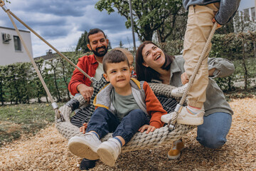 Happy family with two sons playing together on a large rope swing at playground. Parents smiling,...