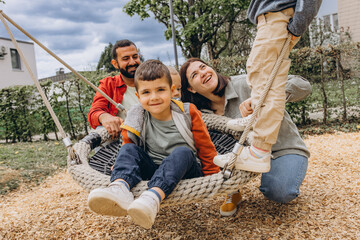 Happy family with two sons playing together on a large rope swing at playground. Parents smiling,...