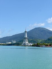 A distinctive white tower stands proudly against a backdrop of lush green mountains, with tranquil turquoise water in the foreground and a bright blue, partly cloudy sky overhead in Langkawi, Malaysia