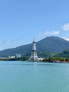 A distinctive white tower stands proudly against a backdrop of lush green mountains, with tranquil turquoise water in the foreground and a bright blue, partly cloudy sky overhead in Langkawi, Malaysia