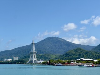 A prominent white tower stands by tranquil blue waters, with a lush green mountain background, a small boat, and scattered buildings under a clear, cloudy sky in Langkawi, Malaysia.