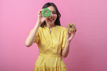 Asian woman in a yellow dress smiling brightly while holding two decorated donuts, one green and...