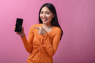 Asian woman with long black hair in orange dress smiling and pointing at smartphone screen with...
