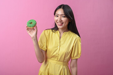 Asian woman in a yellow dress smiling while holding a green-sprinkled donut in front of a pink...