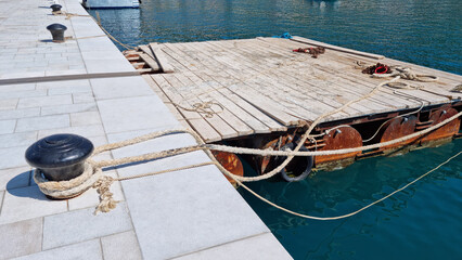 A wooden floating pontoon moored with a rope to a bollard on a paved pier, with blue sea in the...