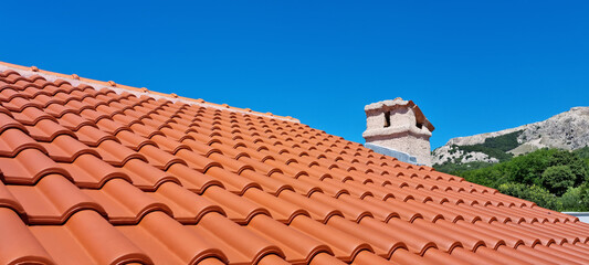 A terracotta tiled roof with a stone chimney under a clear blue sky, with a mountainous landscape in the background © nahhan