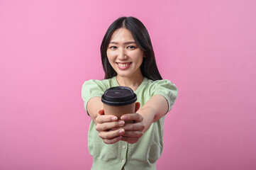 Asian woman smiling while holding a brown takeaway coffee cup with both hands against a solid pink...
