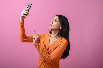 Asian woman with long black hair wearing orange dress pointing upward while holding smartphone in...