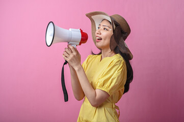 Asian woman in yellow dress and sun hat cheerfully shouting into a megaphone, standing against a...
