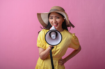 Asian woman in yellow dress and sun hat smiling while speaking into a megaphone against a solid...