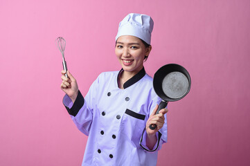 Asian female chef smiling while holding a whisk in one hand and a frying pan in the other, dressed...