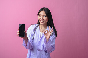 Asian female doctor smiling while showing a smartphone and giving a thumbs up gesture, wearing a...