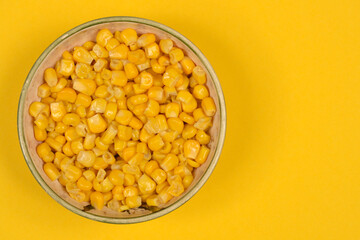 An overhead shot captures a bowl filled with bright yellow corn kernels, positioned on the left side of a vibrant yellow background, with ample copy space