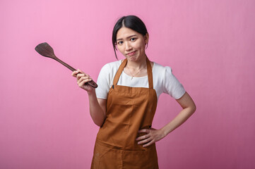 Asian woman in brown apron holding a wooden spatula to her head with a confused expression, posing...