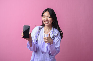 Asian female doctor smiling while showing a smartphone and giving a thumbs up gesture, wearing a...