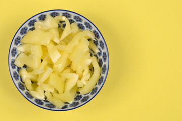 An overhead shot captures a blue and white patterned bowl filled with freshly sliced pale yellow bell peppers, positioned on the left side of a vibrant yellow background with ample copy space