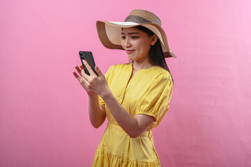 Asian woman in yellow dress and summer hat using smartphone against a pink background in studio...