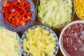 An overhead flat lay showcases a vibrant array of fresh, colorful chopped salad ingredients, including red and yellow bell peppers, shredded lettuce, and beans, presented in various bowls on a wooden 