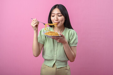 Asian woman in green top eating instant noodles with a fork from a white plate, enjoying her food...