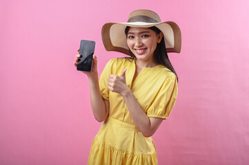 Asian woman in yellow dress and sunhat pointing at smartphone while smiling at the camera, with a...