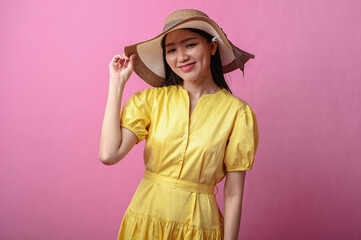 Asian woman in a yellow dress and wide-brim hat smiling softly while touching her hat, posing...