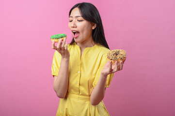 Asian woman in a yellow dress excitedly looking at a green sprinkled donut while holding another...