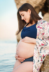 Beautiful young pregnant woman in a bikini sitting on large rocks at the edge of the beach, holding her belly with her hands while looking at the baby she is expecting.