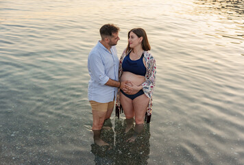 A beautiful couple in love gazing at each other inside the calm waters of the sea, near the shore, both holding the pregnant woman's belly with one hand.