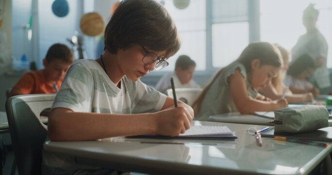 Primary School Children Writing School Test, Doing Tasks in Notebooks. Female Teacher Walking Between Desks, Controlling Group of Diverse Kids During the Exam Process in Modern Classroom. Dolly Shot.